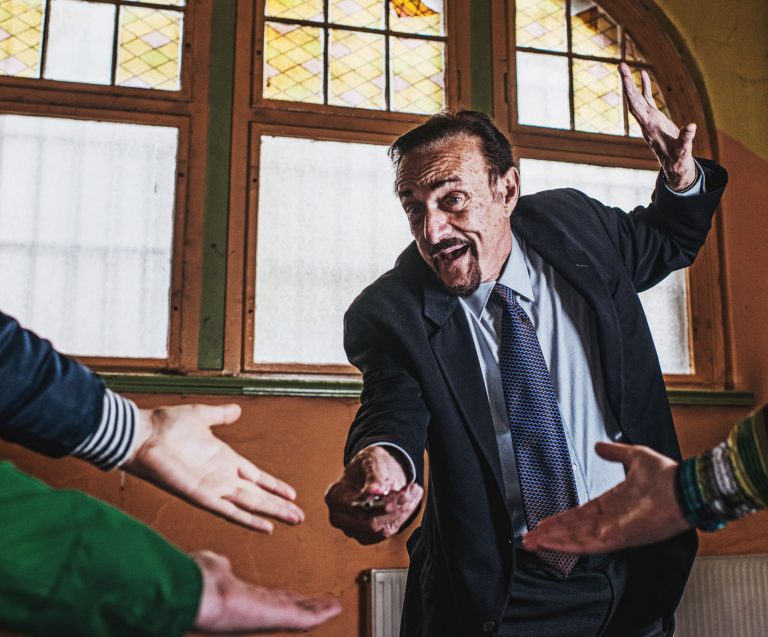 Profesor podaje klucz w kierunku trzech wyciągniętych dłoni. W tle okno z witrażem.