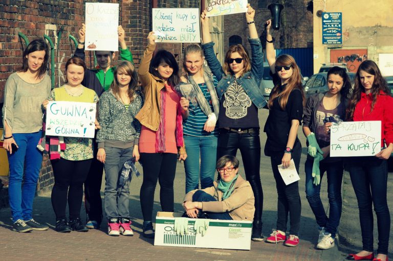 Grupa młodych kobiet stoi na chodniku przed ceglanym budynkiem. Trzymają w rękach kartki reklamujące akcję sprzątania po swoim psie.
