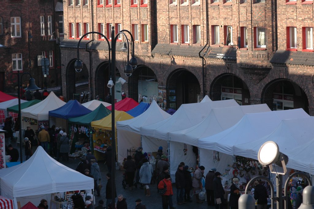 Widok z g&oacute;ry na stoiska wystawowe pod kolorowymi i białymi namiotami. Pomiędzy stoiskami przechodzą ludzie.