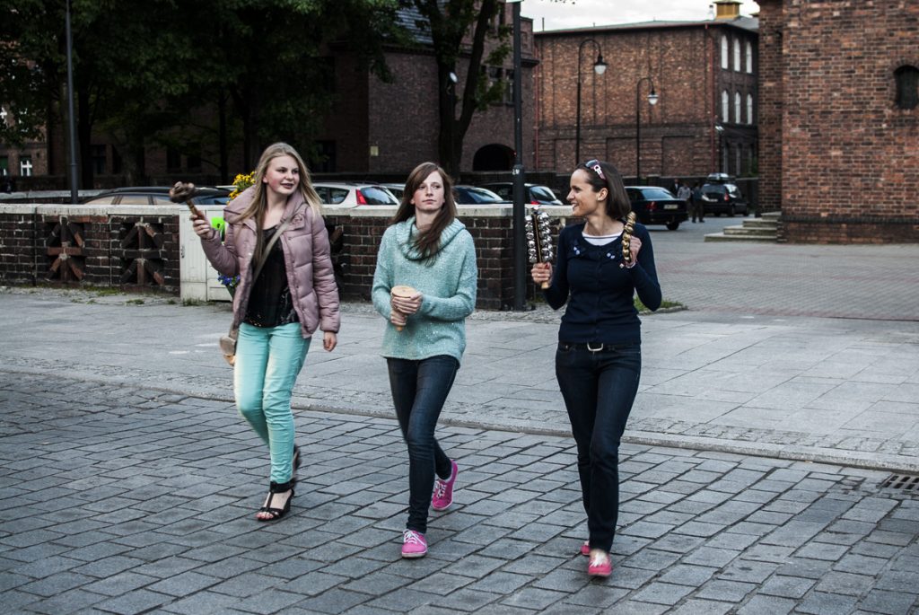 Trzy młode kobiety z instrumentami muzycznymi idą deptakiem. W tle ceglane zabudowania.