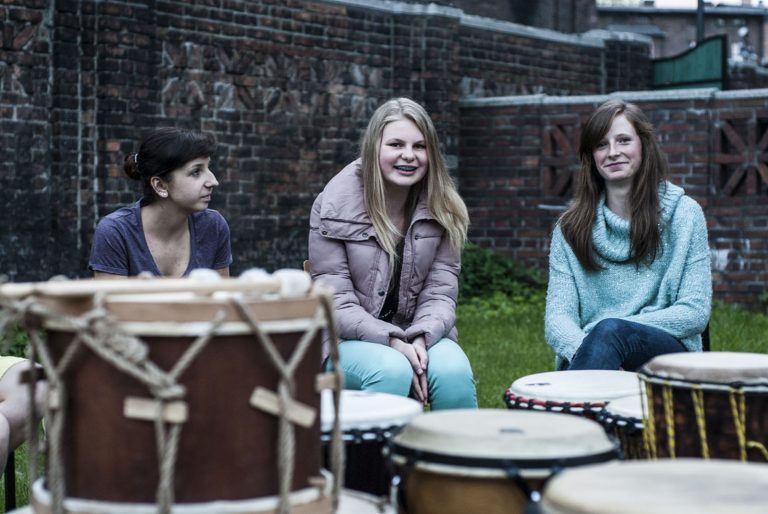 Trzy młode kobiety siedzą na tle ceglanego muru. Przed nimi stoją bongosy.
