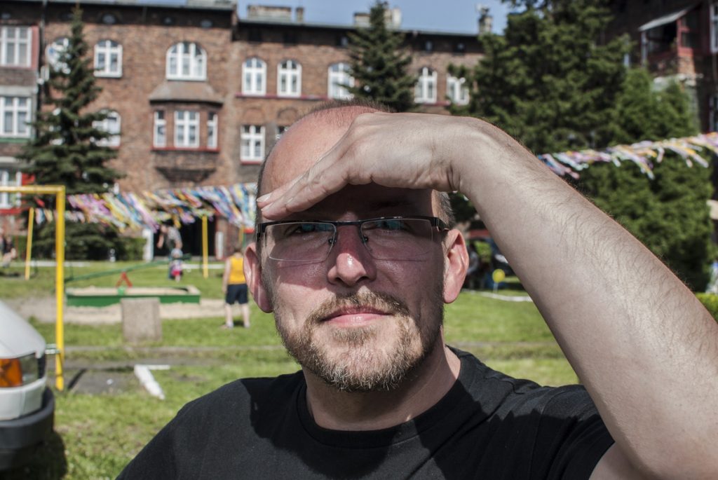 Młody mężczyzna z zarostem i okularach osłania oczy ręką przed słońcem. W tle ceglany budynek.