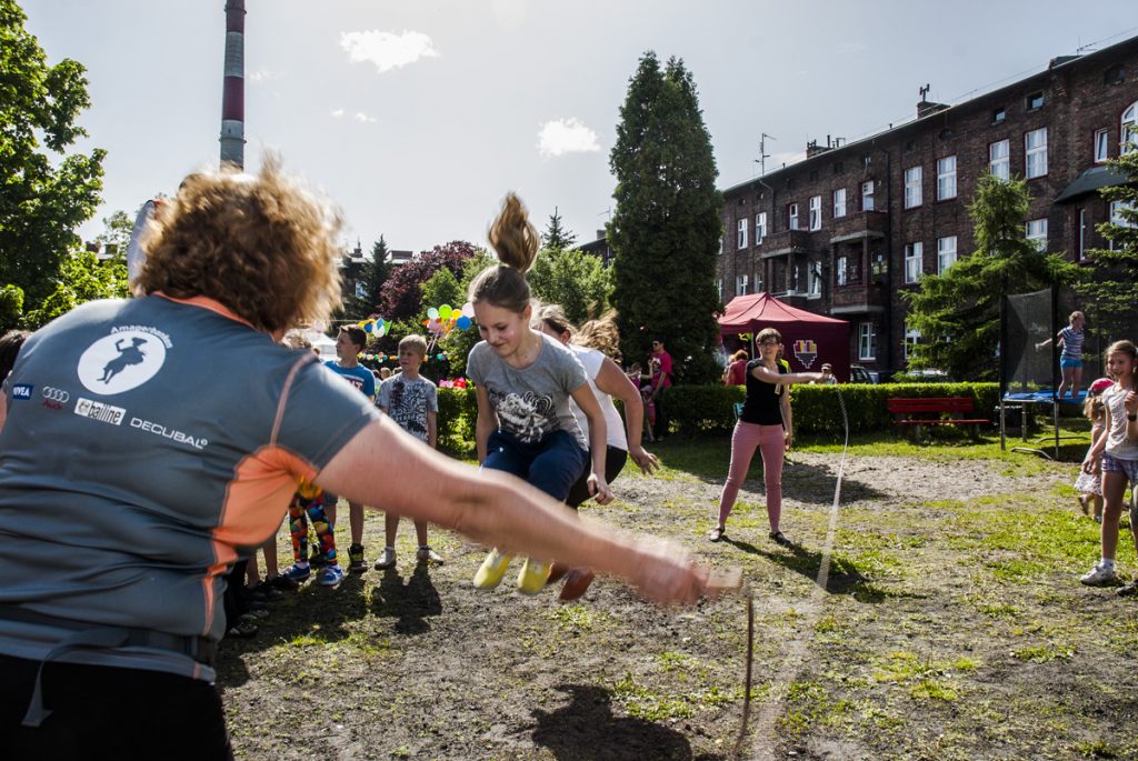Młode dziewczyny skaczą przez skakankę kręconą przez dwie inne osoby. W tle kilkoro dzieci, piknikowy namiot i ceglany budynek.