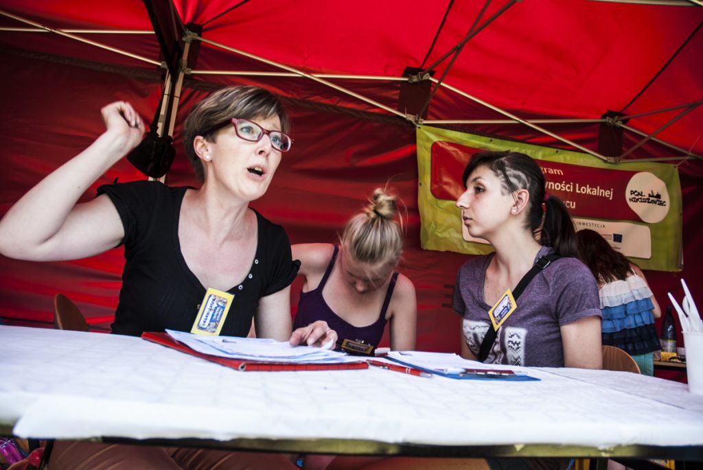 Kobieta w czarnej bluzce i czerwonych okularach z dwiema młodymi dziewczynami siedzi za stołem w czerwonym namiocie.