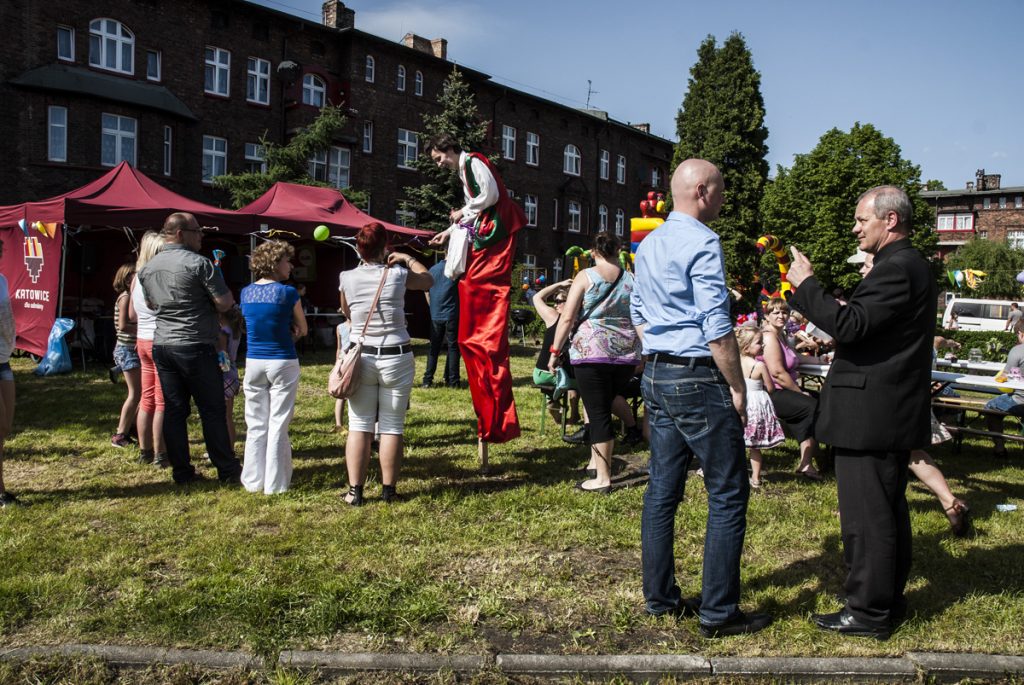 Plenerowy festyn na tle ceglanych budynk&oacute;w. Animator na szczudłach w czerwonym stroju bawi grupę ludzi, a na pierwszym planie rozmawiają dwaj mężczyźni.