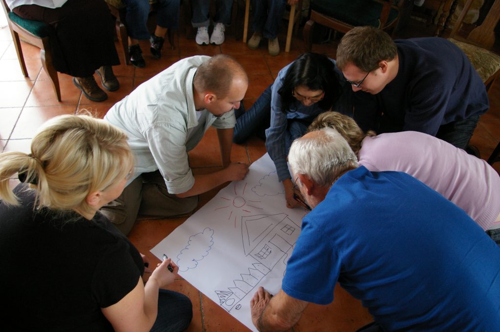 Grupa os&oacute;b klęczy na podłodze z markerami w ręku nad rysunkiem domu.