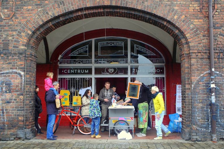 Grupa os&oacute;b przy plenerowym stoisku warsztatowym rozstawionym przed wejściem do Centrum Zimbardo.