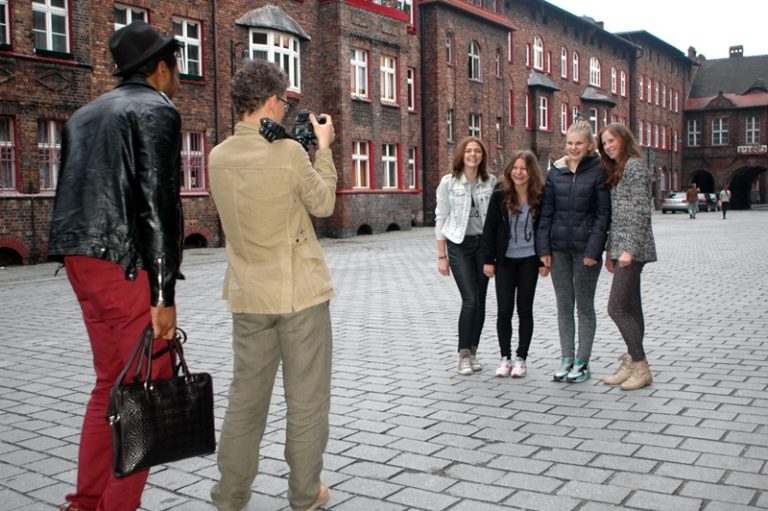 Cztery młode kobiety pozują do zdjęcia na tle kamienic Nikiszowca. Za fotografem stoi mężczyzna w czarnej kurtce, czerwonych spodniach i kapeluszu.