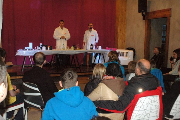 Dwaj mężczyźni w białych fartuchach laboratoryjnych prowadzą pokaz chemiczny przy stole zastawionym odczynnikami, Przed nimi siedzi grupa widz&oacute;w.