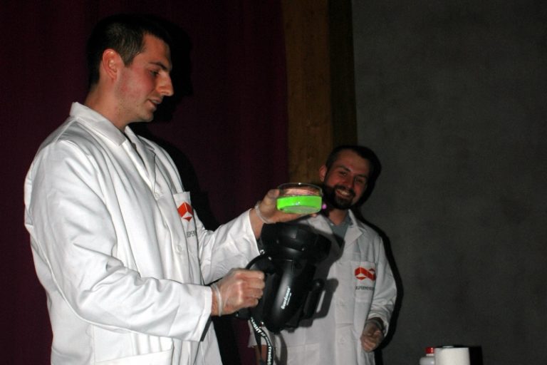 Dw&oacute;ch mężczyzn w białych fartuchach. Jeden z nich trzyma czarny reflektor i szklane naczynie z zieloną cieczą.