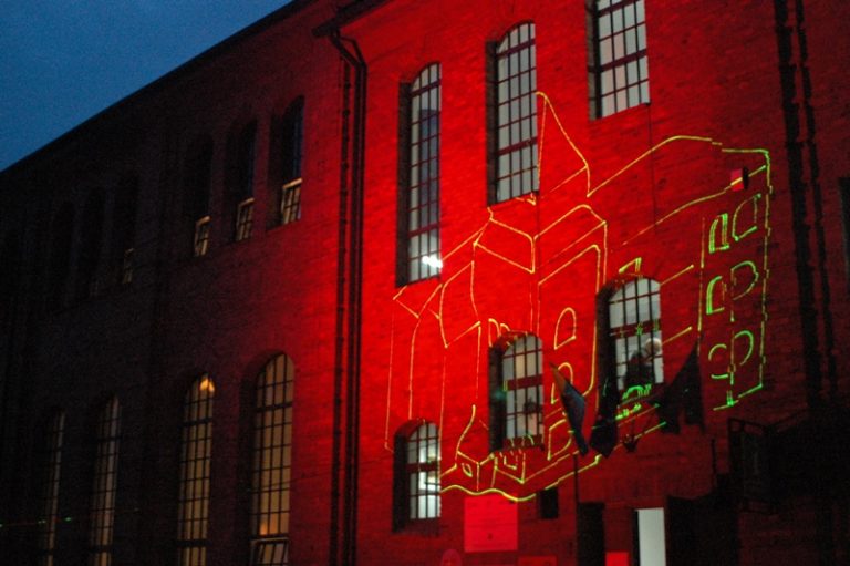 Ceglany budynek po zmroku, podświetlony na czerwono. Na elewacji wyświetlony zielony obrys innego budynku.