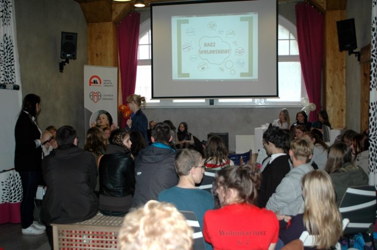 Widok od tyłu na grupę os&oacute;b siedzących przed ekranem rzutnika. Na ekranie wyświetlony jest napis Nasz wolontariat.